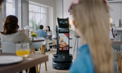 Eine Frau sitzt an einem Tisch, während im Hintergrund der charmante BellaBot Pro-Roboter steht.