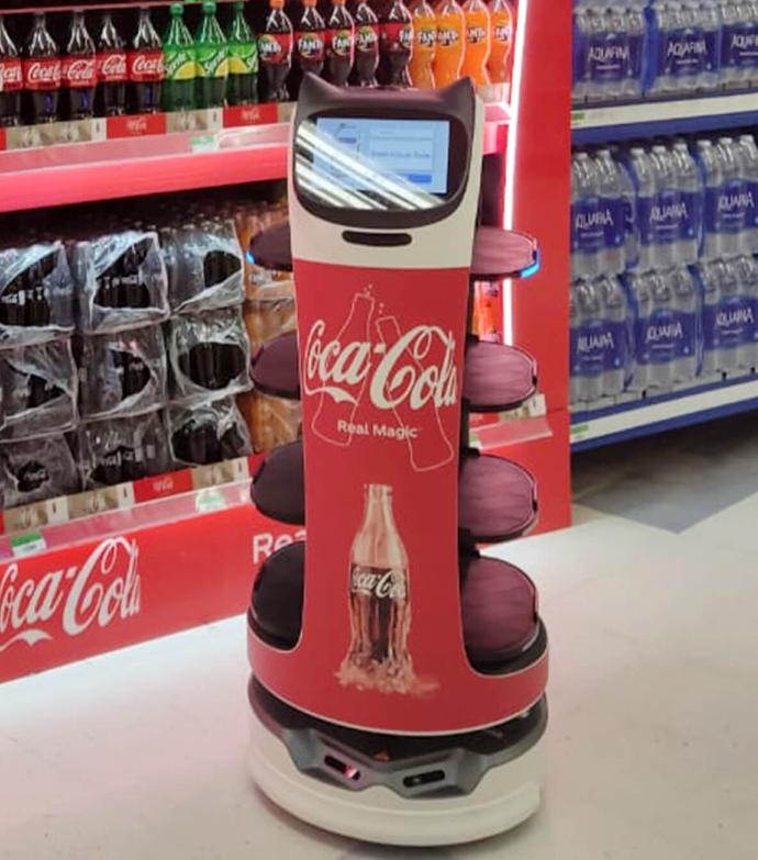 Der Pudu BellaBot-Roboter im Coca-Cola-Design steht vor einem Softdrink-Regal.